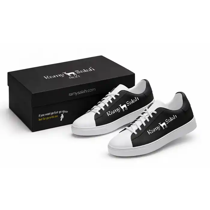 حذاء رياضي أبيض وأسود مع علبة كرتون سوداء مطبوع عليهما شعار رامي صلاح | Black and white sneakers with black shoe box Ramy Salah logo